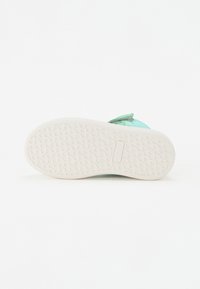 Zapatilla verde con parte superior texturizada, correa ajustable y suela de goma blanca con un patrón ondulado para mayor agarre.