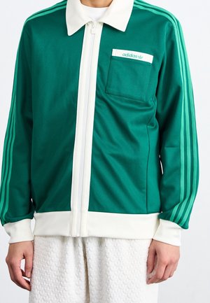 Groene sportjack met witte accenten, volledige ritssluiting aan de voorkant, gestructureerde kraag en een enkele borstzak. Bevat drie bijpassende strepen op de mouwen.