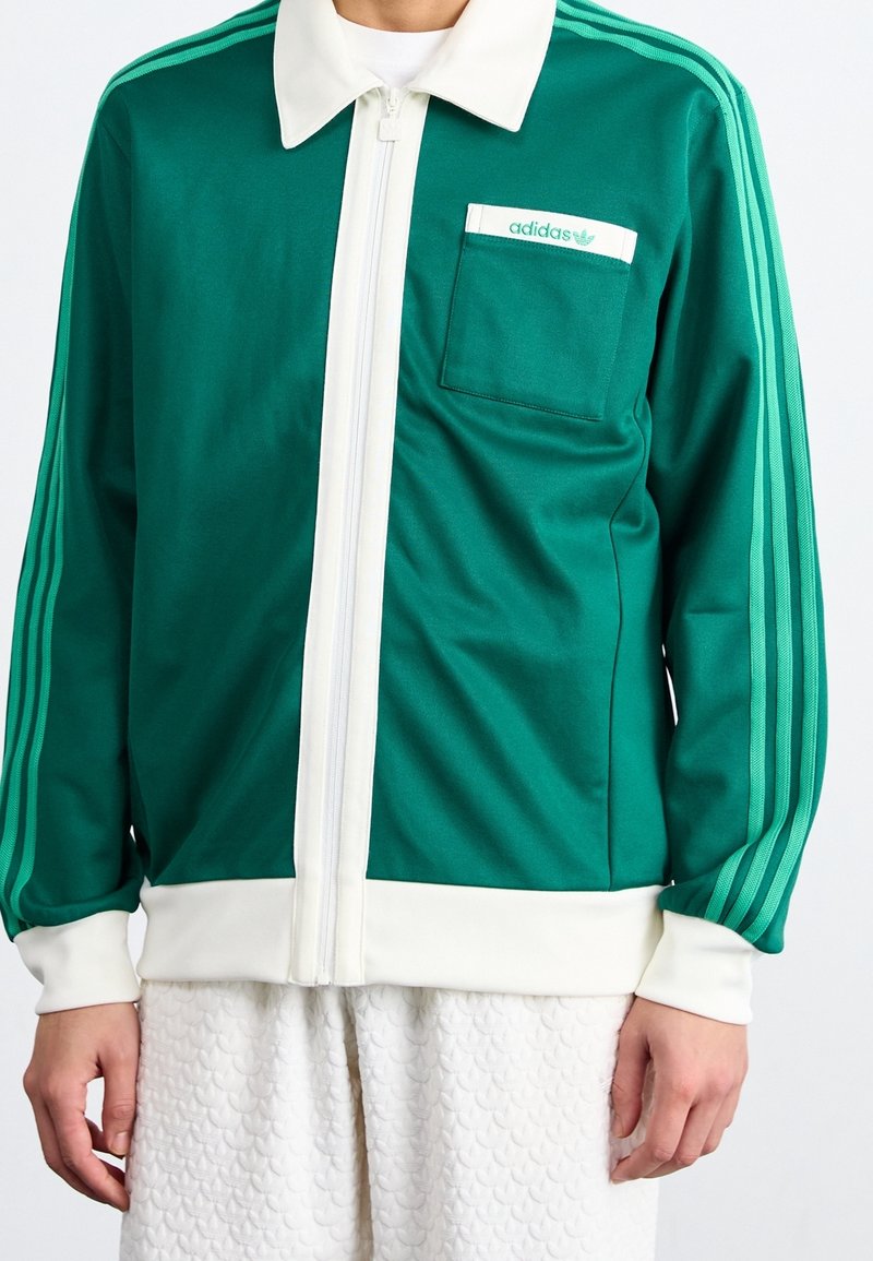 Veste de sport verte avec des accents blancs, fermeture éclair sur le devant, col structuré et une poche poitrine. Présente trois rayures assorties sur les manches.