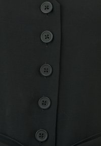 Top in tessuto nero con sei bottoni neri verticali, caratterizzato da una superficie testurizzata e un design strutturato incentrato sulla funzionalità.