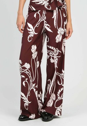 Pantalon marron à jambes larges avec de grands motifs floraux blancs, porté avec des chaussures noires brillantes, raccourci à la taille et aux chevilles.