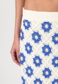 Heklet skjørt med hvit base og blå blomster mønster samt beige detaljer, som viser et teksturert blonderdesign.