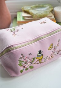 Lichtroze etui met rits, geborduurde vogel, vlinder, bij en een takje van kersenbloesem, geplaatst op een wit oppervlak naast boeken en een kaars.