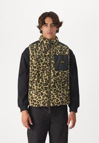 Fleeceová vesta s leopardím vzorom v béžovej a čiernej farbe, má predný zips a čiernu vrecko s žltým logom. Čierne rukávy a robustná textura.