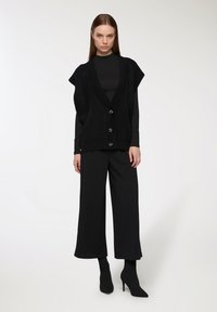 Gilet senza maniche nero a maglia con scollatura a V e bottoni frontali, indossato sopra una maglietta a maniche lunghe nera, abbinato a pantaloni neri larghi e calze con tacco.