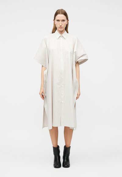 Woman wearing a loose, knee-length, light beige button-up dress with short wide sleeves and black ankle boots against a white background.