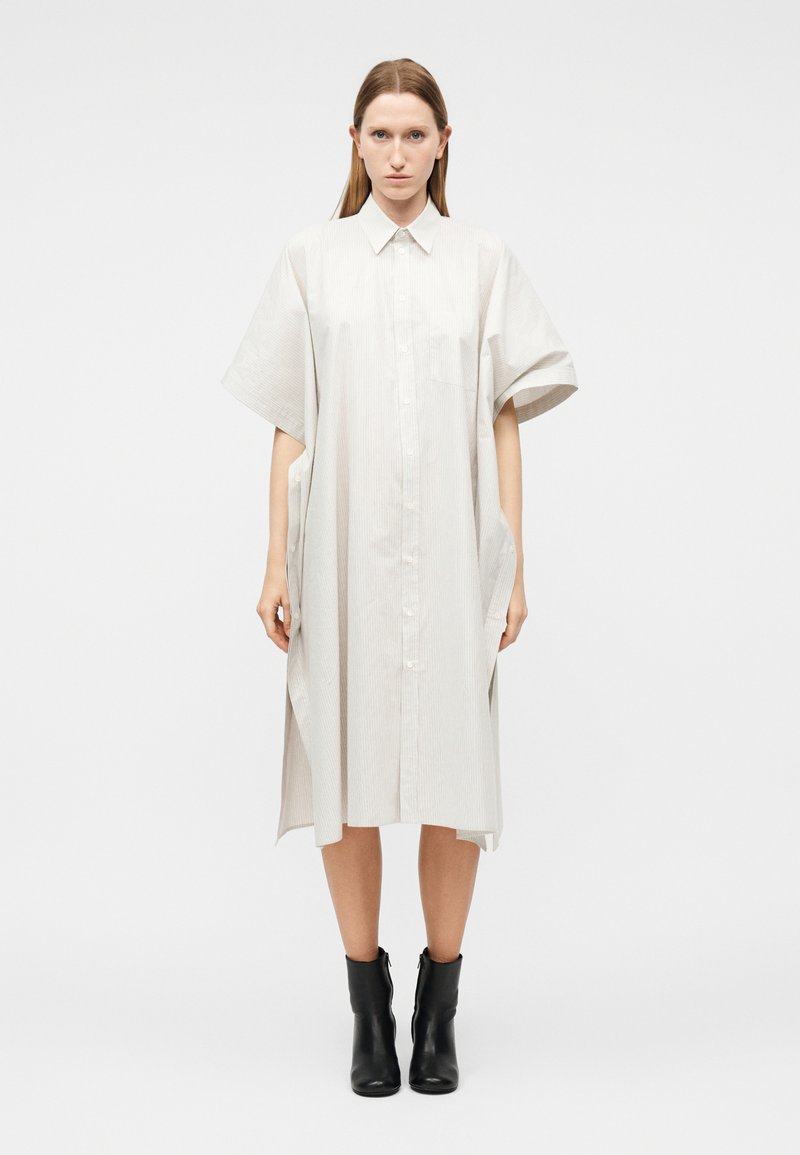 Femme portant une robe boutonnée ample, longueur au genou, beige clair, avec des manches courtes larges et des bottines noires, sur fond blanc.