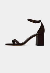 Καφέ δερμάτινο σανδάλι με χοντρό τακούνι, με φαρδιά λουράκι πάνω από τα δάχτυλα και λεπτό λουρί στον αστράγαλο με χρυσή ρυθμιζόμενη πόρπη.