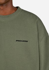 Ingrandimento di una persona che indossa un felpa girocollo verde oliva con il testo nero "PEGADOR" ricamato sul petto.