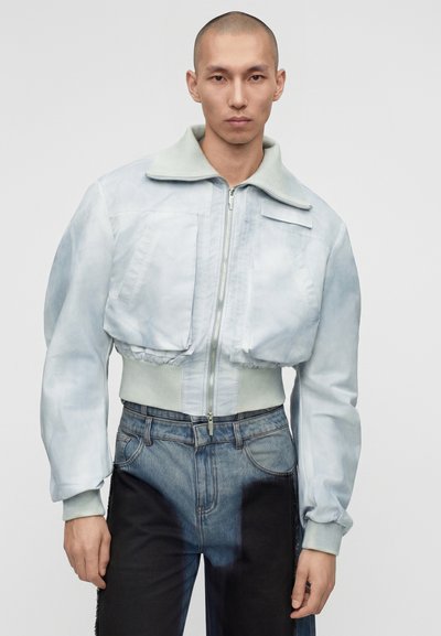 Άνδρας με κοντό ανοιχτό μπλε bomber μπουφάν με μεγάλες μπροστινές τσέπες και δίχρωμα τζιν, στέκεται μπροστά σε απλό φόντο.