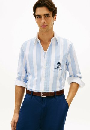 Mand iført en lyseblå og hvid skjorte med lodrette striber og op rullede ærmer samt marineblå bukser, stående med den ene hånd i lommen.