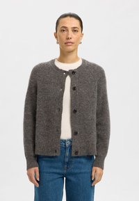 Šedý kardigan z měkké, texturované látky s kulatým výstřihem, zapínáním na knoflíky vpředu a dlouhými rukávy; doplněný o světlé tričko a džíny.