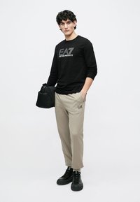 Čierna dlhá košeľa s logom "EA7 Emporio Armani", béžové nohavice a čierne tenisky. Model drží čiernu tašku a stojí na svetlom pozadí.