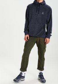 Marinblå hoodie med snörning och frontlogotyp; olivgröna cargo-byxor med klafffickor; grå sneakers med vita detaljer och mönstrade strumpor.