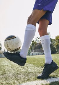 Svarta fotbollsskor med texturerat grepp, vita knähöga strumpor och en svart-vit fotboll på grön gräsyta, med en suddig bakgrund.