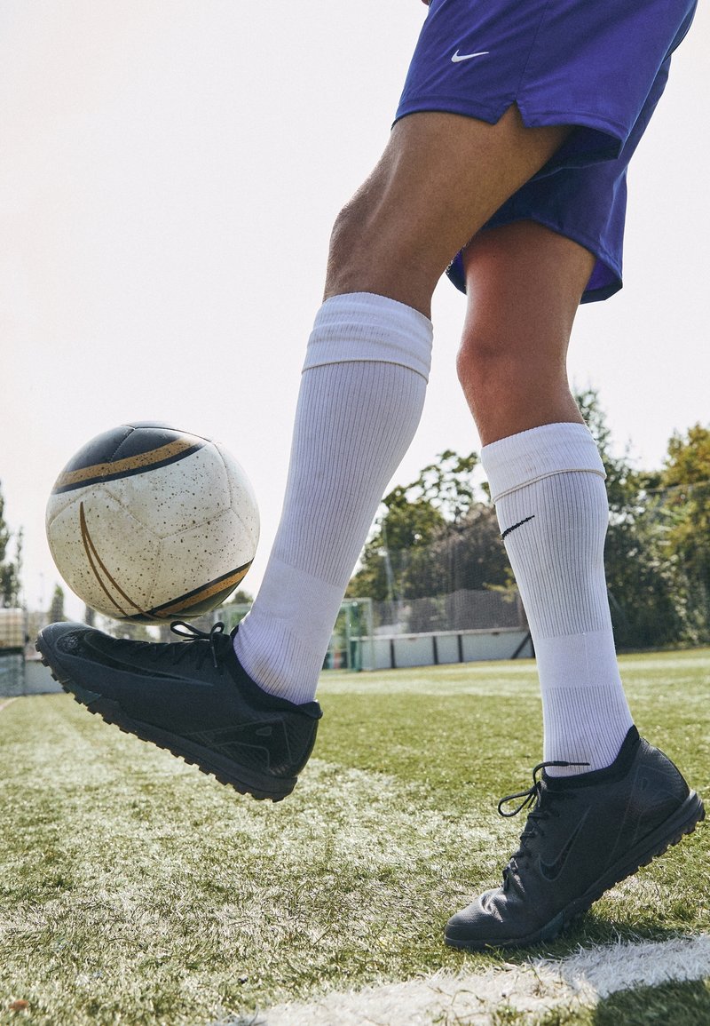 Svarta fotbollsskor med texturerat grepp, vita knähöga strumpor och en svart-vit fotboll på grön gräsyta, med en suddig bakgrund.