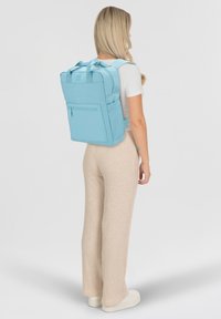 Zaino azzurro chiaro con spallacci imbottiti, tasca frontale con zip e manici superiori. Texture liscia e forma rettangolare, adatto per trasportare l'essenziale.