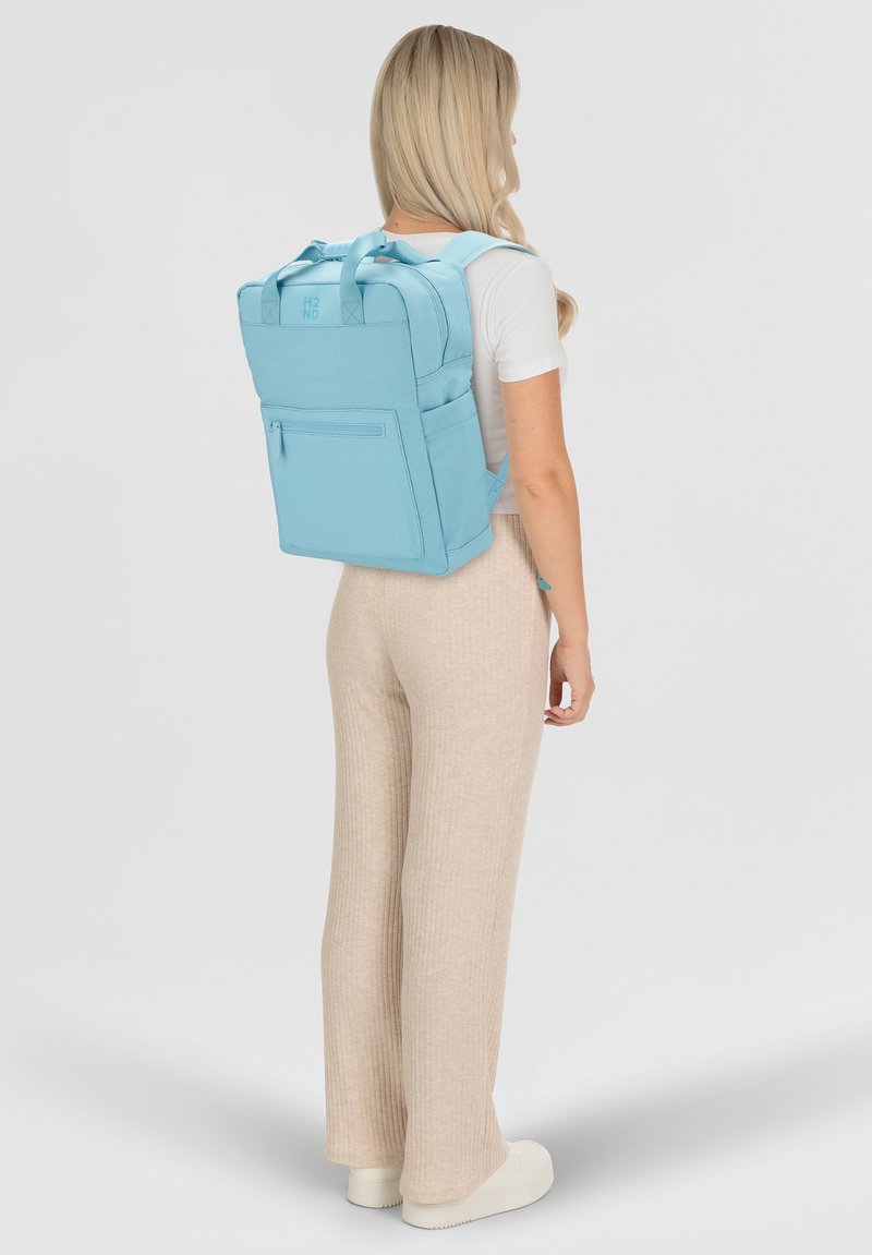 Zaino azzurro chiaro con spallacci imbottiti, tasca frontale con zip e manici superiori. Texture liscia e forma rettangolare, adatto per trasportare l'essenziale.