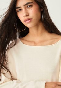 Pull en maille beige clair avec un col large, texture lisse et manches longues. Le modèle porte de grandes boucles d'oreilles et a les cheveux longs et noirs.
