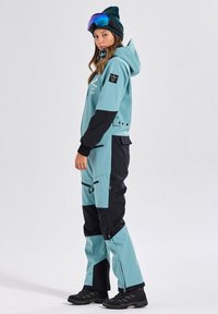 Turquoise and black ski suit with a hood, zip pockets, and logo accents. Worn with a beanie and goggles, featuring a slim, fitted design.