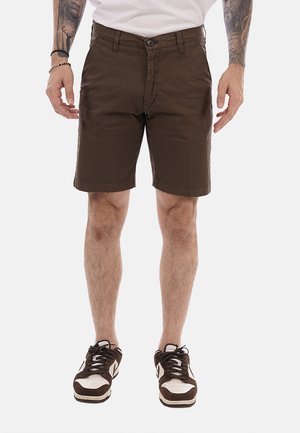 Uomo che indossa pantaloncini marroni al ginocchio con bottone e cerniera, scarpe da ginnastica bianche e tatuaggi visibili sulle braccia, in piedi con le braccia rilassate.