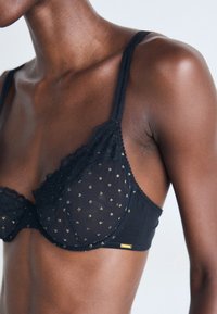 Soutien-gorge en dentelle noire avec des pois dorés porté par une personne à la peau foncée, montrant le haut du torse et l'épaule sur un fond uni.