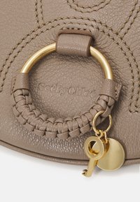 Bolso de mano de cuero marrón con un asa trenzada, herrajes en tono dorado y un detalle de llavero con una etiqueta circular y un acento de llave.