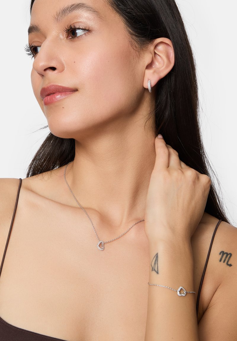 Smycken i silver med hjärtformat hänge, en halskedja och armband, båda prydda med små ädelstenar, minimalistisk design, slät yta.