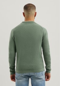 Green textured knit sweater with long sleeves and a polo collar, paired with light blue jeans. Back view showcases stitching details.