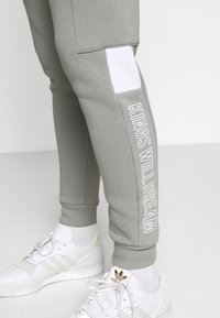 Graue Jogginghose mit einem weißen Einsatz an der Seite und dem weißen Schriftzug "KINGS WILL DREAM"; kombiniert mit weißen Sportschuhen.