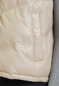 Beige leren jas met een gestructureerd, doorgestikt ontwerp, een zijzak en zichtbare stiksels. De binnenkant heeft een grijze, geribbelde stof.