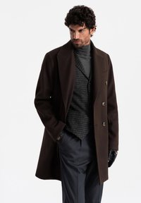 Brown wool overcoat with large lapels and double-breasted buttons over a gray turtleneck and plaid vest; paired with gray trousers and black gloves.