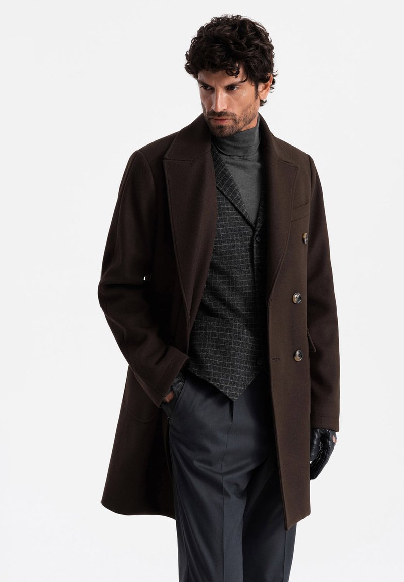 Brown wool overcoat with large lapels and double-breasted buttons over a gray turtleneck and plaid vest; paired with gray trousers and black gloves.