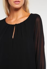 Czarna bluzka z okrągłym dekoltem, z plisowanym przodem i przezroczystymi długimi rękawami, z małym otworem w kształcie kluczyka na dekolcie.