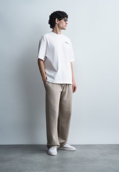 Άνδρας με σγουρά μαλλιά και γυαλιά ηλίου στέκεται μπροστά σε ένα απλό τοίχο φορώντας μια λευκή φαρδιά μπλούζα, μπεζ φαρδιά παντελόνια και λευκά αθλητικά παπούτσια.