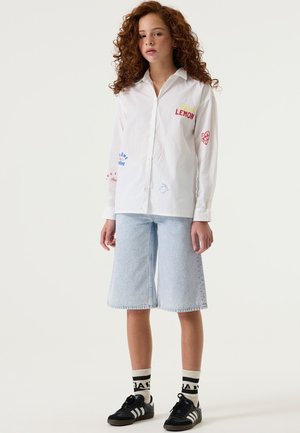 Camisa blanca de algodón con botones, con texto y gráficos impresos en colores; combinada con culottes de mezclilla azul claro y zapatillas negras con calcetines a rayas.