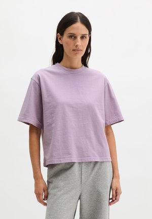 Kobieta o długich ciemnych włosach, ubrana w luźny jasnofioletowy T-shirt z krótkim rękawem i jasnoszare spodnie, stojąca na tle jednolitego białego tła.