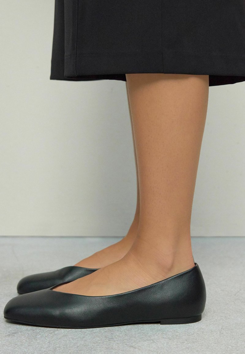 Musta nahkainen ballerinatyyppinen kengät, joissa on pyöreä kärki ja minimalistiinen muotoilu. Siro pinta ja tasapohja, asetettuna vaaleanharmaalle pinnalle.