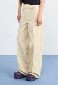 A person stands wearing beige wide-leg trousers and purple clogs, with a minimalist background and light blue floor.