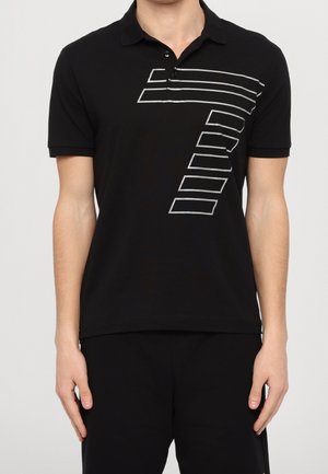 Mann trägt ein schwarzes, kurzärmeliges Poloshirt mit einem geometrischen weißen Umrissmuster auf der Brust, kombiniert mit einer schwarzen Hose.