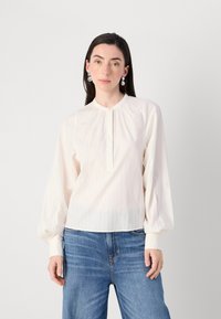 Il modello indossa una camicia a maniche lunghe di colore chiaro con colletto alto e bottoni, abbinata a jeans blu, davanti a uno sfondo semplice.