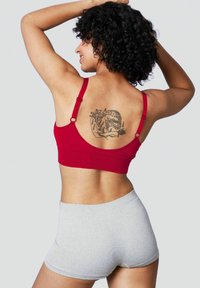 Bralette rossa con spalline regolabili, tessuto liscio e schiena bassa, abbinato a pantaloni corti grigi con texture. Tatuaggi visibili sulla schiena.