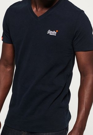 Mand iført en ensfarvet mørkeblå V-hals T-shirt med et lille hvidt og orange logo på venstre bryst og en lille mærkat på venstre ærme.