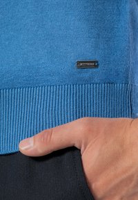 Niebieski sweter z dzianiny z prążkowanym dołem i mankietami; posiada małą, metaliczną metkę wszytą w materiał. Zbliżenie pokazuje rękę w kieszeni.