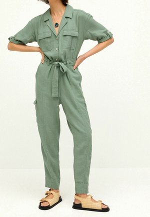 Combi-pantalon en lin vert avec manches retroussées, deux poches poitrine et une ceinture nouée à la taille. Associée à des sandales camel et un collier pendentif sombre.