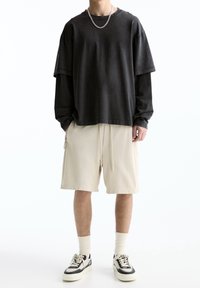 Person wearing layered black long-sleeve and short-sleeve shirts, beige knee-length shorts, white socks, black and white sneakers, and a silver chain.