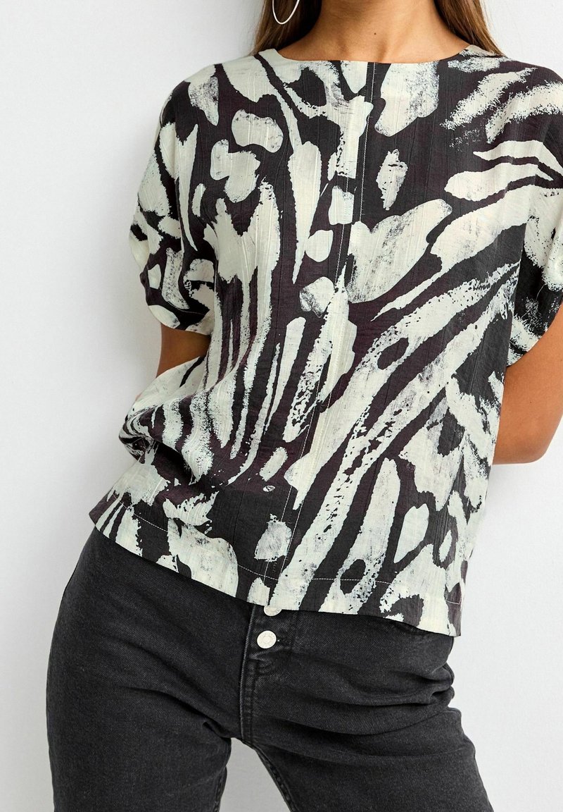 Femme portant un chemisier à manches courtes à motif abstrait noir et blanc, associé à un jean noir taille haute avec des détails à boutons.