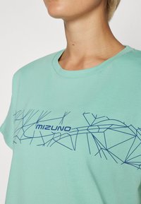 Mintgrønn bomull T-skjorte med rund hals, med et blått geometrisk linjemønster og ordet "MIZUNO" trykt under grafikken.