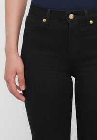 Jeans in denim nero con una vestibilità slim, dotati di bottoni dorati, design a cinque tasche e una texture liscia. Mano appoggiata sulla coscia.