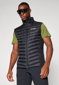 Black insulated vest with a quilted design and front zipper, paired with a short-sleeve green shirt. Features a logo on the chest.